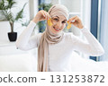 Young Muslim woman focused on skincare routine using gold patches on eye area, showcasing beauty and self-care. Relaxing indoor setting, wearing beige hijab and white top, maintaining wellness. 131253872