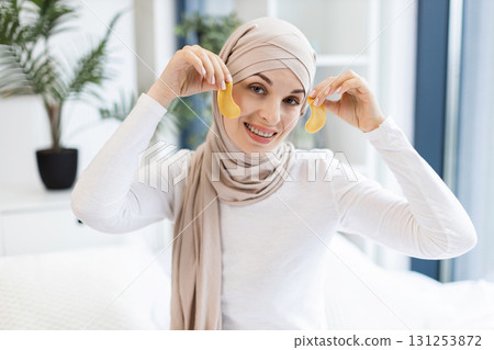 Young Muslim woman focused on skincare routine using gold patches on eye area, showcasing beauty and self-care. Relaxing indoor setting, wearing beige hijab and white top, maintaining wellness. Young Muslim woman focused on skincare routine using gold patches on eye area, showcasing beauty and self-care. Relaxing indoor setting, wearing beige hijab and white top, maintaining wellness. 131253872