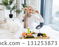 Young adult Muslim woman performing skincare routine using gold hydrogel eye patches, surrounded by fresh fruits and cosmetics, sitting on bed in cozy home environment, promoting wellness and beauty. 131253873