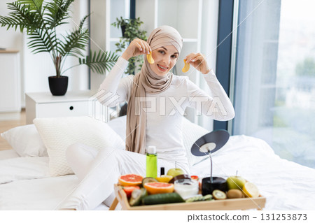 Young adult Muslim woman performing skincare routine using gold hydrogel eye patches, surrounded by fresh fruits and cosmetics, sitting on bed in cozy home environment, promoting wellness and beauty. Young adult Muslim woman performing skincare routine using gold hydrogel eye patches, surrounded by fresh fruits and cosmetics, sitting on bed in cozy home environment, promoting wellness and beauty. 131253873