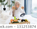 Young Muslim woman on bed applies gold hydrogel patches for eye care. Scene highlights skincare, beauty, self-care routines with natural ingredients. 131253874