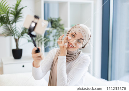 Young adult Muslim woman with hijab applying facial cream while using smartphone for vlogging. Captures concept of modern self-care and engagement through social media. 131253876