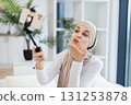 Young adult Muslim woman with hijab applying facial cream while using smartphone for vlogging. Captures concept of modern self-care and engagement through social media. 131253878