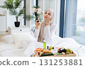 Smiling young Muslim woman applies skincare cream while recording vlog in bright bedroom. Relaxed atmosphere with natural light, modern decor, fresh fruits. Ideal homecare, beauty, content creation. 131253881