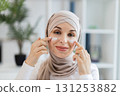 Young Muslim woman in beige hijab applying facial cream on cheeks, promoting skincare. Cream application emphasizes self-care, wellness, personal beauty routine in serene environment. 131253882