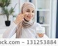 Smiling adult Muslim woman wearing hijab applying moisturizer cream on facial skin indoors. Promotes skincare, self-care, beauty regime, and wellness. 131253884