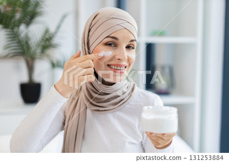 Smiling adult Muslim woman wearing hijab applying moisturizer cream on facial skin indoors. Promotes skincare, self-care, beauty regime, and wellness. 131253884