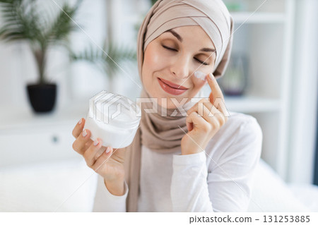 Smiling adult Muslim woman wearing hijab applying moisturizer cream on facial skin indoors. Promotes skincare, self-care, beauty regime, and wellness. Smiling adult Muslim woman wearing hijab applying moisturizer cream on facial skin indoors. Promotes skincare, self-care, beauty regime, and wellness. 131253885