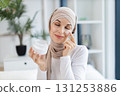 Young adult Muslim woman in hijab skincare routine at home with facial cream. Expresses happiness and simple beauty self-care. Bright indoor atmosphere showcasing plant in stylish decor 131253886