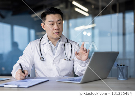 A young Asian male doctor sits at a desk in a clinic, conducts an online consultation on a laptop, makes notes in a notebook. 131255494