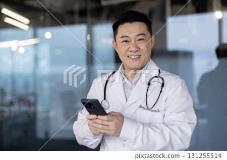 Portrait of a smiling young Asian doctor in a white coat standing in the hospital lobby, holding a mobile phone and looking at the camera. 131255514