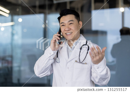An asian male doctor, wearing a lab coat and a stethoscope, is on a phone call inside the clinic. He gestures with his hand while talking. An asian male doctor, wearing a lab coat and a stethoscope, is on a phone call inside the clinic. He gestures with his hand while talking. 131255517