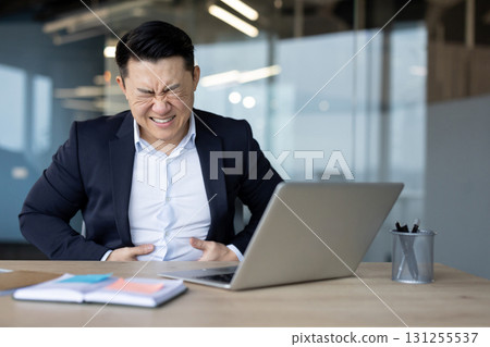 A young Asian man, grimacing in pain, sits at his workplace and holds his stomach with his hands. A young Asian man, grimacing in pain, sits at his workplace and holds his stomach with his hands. 131255537