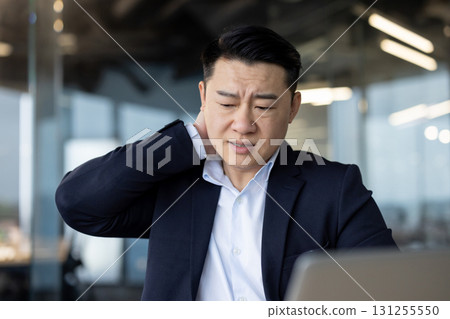 Close-up photo of a young Asian businessman sitting tiredly at his workplace, holding his neck, massaging it with his hand, feeling severe pain and tension. Close-up photo of a young Asian businessman sitting tiredly at his workplace, holding his neck, massaging it with his hand, feeling severe pain and tension. 131255550