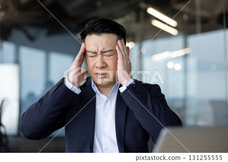 Close-up photo of a young tired Asian businessman in the office, standing holding his head in his hands, feeling severe pain. Close-up photo of a young tired Asian businessman in the office, standing holding his head in his hands, feeling severe pain. 131255555