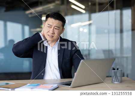 A young Asian businessman, twisted in pain, sits exhausted at his desk in the office and massages his neck with his hand. 131255558