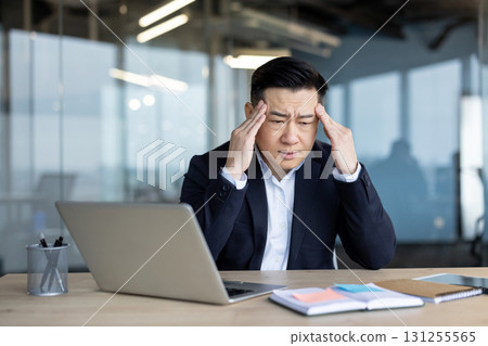 Tired young Asian businessman sitting at the desk in the office, holding his head thoughtfully, feeling strong pressure and pain. Tired young Asian businessman sitting at the desk in the office, holding his head thoughtfully, feeling strong pressure and pain. 131255565
