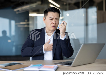 An asian businessman in a suit uses an inhaler while working on a laptop in the office, managing his health. 131255577