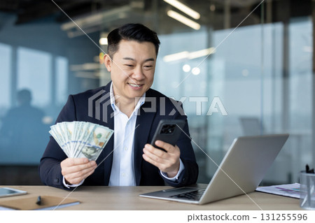 Asian young male businessman sitting in the office at a desk with a notebook, holding a mobile phone and a fan of dollar bills. 131255596