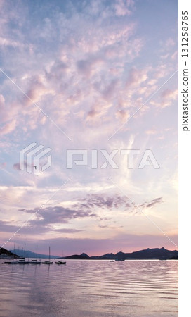 A peaceful coastal sunset with soft pink and purple clouds reflected on the calm water, with a silhouette of distant hills and a line of sailboats. 131258765