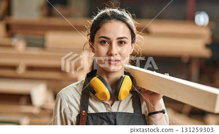 Crafting with confidence, woman in workshop carries wood while wearing safety gear in a busy lumber environment 131259303