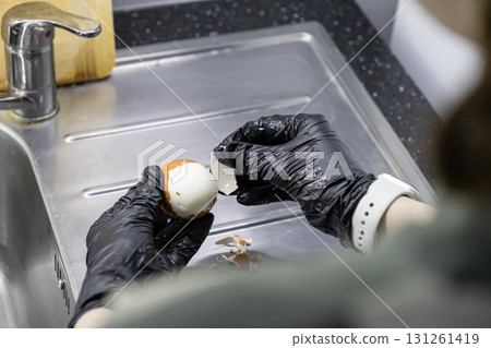 Hands in black protective gloves meticulously peel a freshly hard-boiled chicken egg over a clean stainless steel kitchen sink, ensuring safe food preparation 131261419