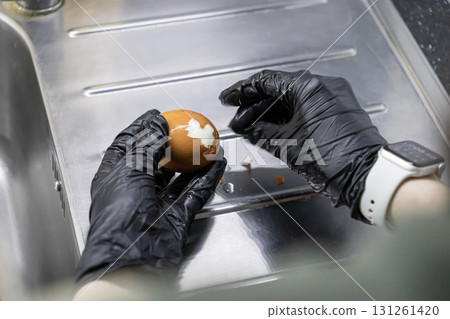 Hands in black gloves meticulously peeling a brown hard-boiled egg over a clean stainless steel kitchen sink, ensuring safe food handling and preparation hygiene Hands in black gloves meticulously peeling a brown hard-boiled egg over a clean stainless steel kitchen sink, ensuring safe food handling and preparation hygiene 131261420