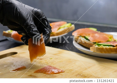 Chef's gloved hand expertly arranges fresh salmon fillet on a wooden board, crafting gourmet sandwiches, highlighting healthy food preparation and culinary skill 131261421