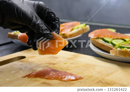 Professional chef's gloved hand carefully placing fresh smoked salmon onto a toast with cream cheese and cucumber, preparing healthy gourmet appetizer snacks Professional chef's gloved hand carefully placing fresh smoked salmon onto a toast with cream cheese and cucumber, preparing healthy gourmet appetizer snacks 131261422