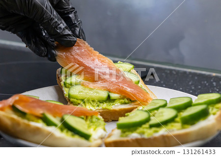 Gloved hand carefully placing fresh salmon slices onto toasted bread with creamy avocado spread and crisp cucumber for a healthy gourmet snack Gloved hand carefully placing fresh salmon slices onto toasted bread with creamy avocado spread and crisp cucumber for a healthy gourmet snack 131261433