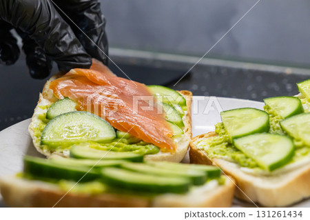 Gloved hand placing smoked salmon on a fresh avocado and cream cheese toast with cucumber slices, perfect for a healthy breakfast Gloved hand placing smoked salmon on a fresh avocado and cream cheese toast with cucumber slices, perfect for a healthy breakfast 131261434