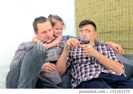 Gay family with daughter eat tangerine and drink wine together in living room. Gay family with daughter eat tangerine and drink wine together in living room. 131262370