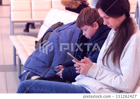 Woman with her son browsing on mobile phones in airport hall waiting for flight. Side view. Woman with her son browsing on mobile phones in airport hall waiting for flight. Side view. 131262427
