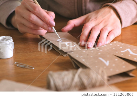 Woman makes christmas advent calendar for kids. Writes a number on the bag by brush and white paint. Close-up hands. 131263005