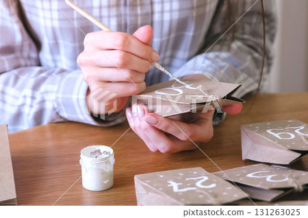 Woman makes christmas advent calendar for kids. Writes a number on the bag by brush and white paint. Close-up hands. Woman makes christmas advent calendar for kids. Writes a number on the bag by brush and white paint. Close-up hands. 131263025
