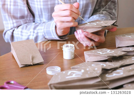 Woman makes christmas advent calendar for kids. Writes a number on the bag by brush and white paint. Close-up hands. 131263028