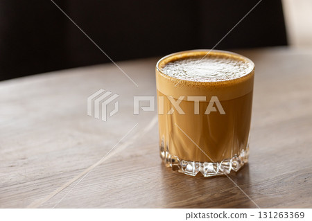 Flat white. A creamy coffee in a clear glass rests on a wooden table, close up 131263369