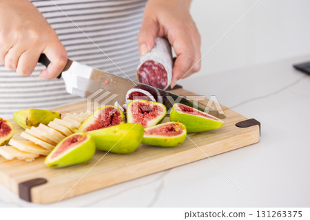 A person slices fresh green figs, dry-cured sausage and cheese on a cutting board 131263375