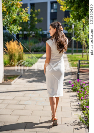 Young woman wearing a white midi dress walking outdoors in Old Money style. Concept of timeless fashion, graceful femininity and understated elegance in modern lifestyle. Young woman wearing a white midi dress walking outdoors in Old Money style. Concept of timeless fashion, graceful femininity and understated elegance in modern lifestyle. 131263489
