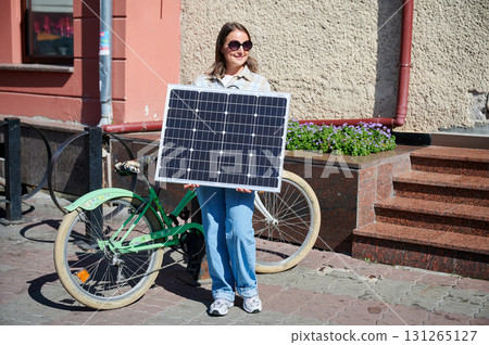 Smiling woman holding solar panel, standing beside green bicycle. Happy female in sunglasses and plaid shirt, enjoying sunny urban day. Smiling woman holding solar panel, standing beside green bicycle. Happy female in sunglasses and plaid shirt, enjoying sunny urban day. 131265127