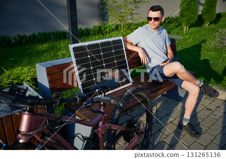 Man cyclist using solar panel for charging electric mountain bike outdoors. Guy in sunglasses sits on bench, holding phone, casually dressed in t-shirt, shorts, enjoying sunny outdoor setting. 131265136