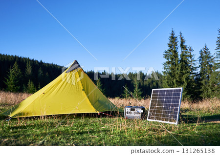 Tourist tent with solar panel and portable power station nearby in grassy field. Scene surrounded by rolling hills, tall pine trees under clear blue sky, showcasing eco-friendly camping setup. Tourist tent with solar panel and portable power station nearby in grassy field. Scene surrounded by rolling hills, tall pine trees under clear blue sky, showcasing eco-friendly camping setup. 131265138