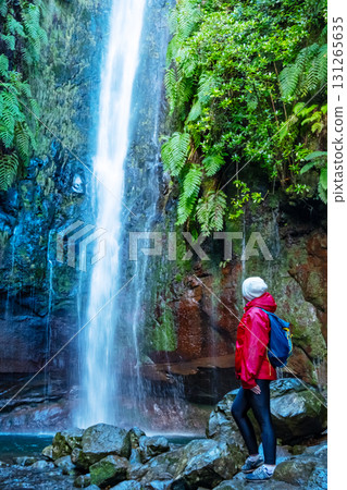 Hiker in red jacket exploring a scenic levada trail with waterfall surrounded by twisted forest trees in Madeira. The nature walk provides a peaceful and adventurous experience Hiker in red jacket exploring a scenic levada trail with waterfall surrounded by twisted forest trees in Madeira. The nature walk provides a peaceful and adventurous experience 131265635