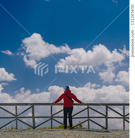 Individual stands on a balcony in a red jacket, gazing towards a partially cloudy sky, embodying a sense of contemplation. Wooden railing frames the expansive view. 131265650