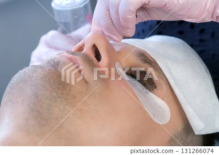Beautician wiping black paint from man's eyelashes on laminating lash procedure. 131266074