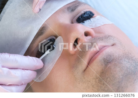 Beautician wiping black paint from man's eyelashes on laminating lash procedure. Beautician wiping black paint from man's eyelashes on laminating lash procedure. 131266076