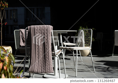 Cozy outdoor setting with modern table and minimalist white chairs. Soft knit blanket is draped over chair, creating welcoming and stylish atmosphere under warm sunlight Cozy outdoor setting with modern table and minimalist white chairs. Soft knit blanket is draped over chair, creating welcoming and stylish atmosphere under warm sunlight 131268487