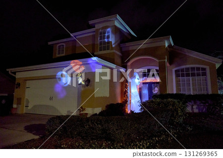 A suburban house shows a glowing ghost inflatable by the entrance and a colorful skull-and-moon light projection on the garage wall. Nighttime decorations create a spooky, festive holiday display. 131269365