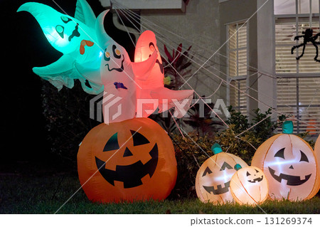 Three glowing ghosts rise from a jack-o-lantern inflatable beside spider webs at night. Bright colors and cheerful faces create a festive, spooky front-yard Halloween scene. 131269374