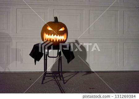 A carved jack-o-lantern pumpkin with sharp teeth glows on a small table in front of a white garage door at night. Minimal Halloween decor with strong shadows, orange light, and spooky mood. 131269384